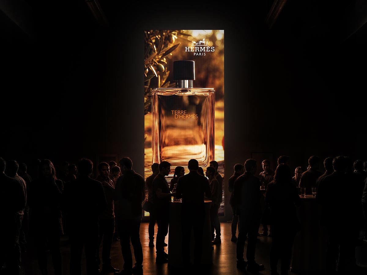 A tall illuminated screen in a darkened event space, guests silhouetted in front of it.